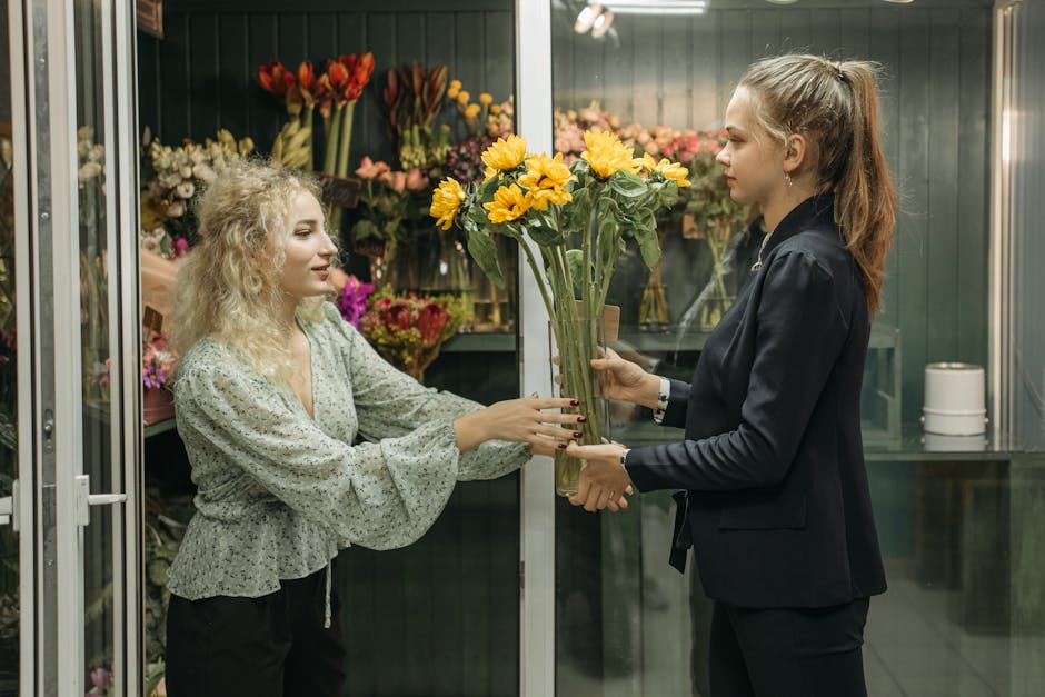 A floral assistant with light brown hair tied back in a ponytail, dressed in a black blazer, hands a tall bouquet of bright yellow sunflowers and smaller pink and purple flowers to a customer with cur
