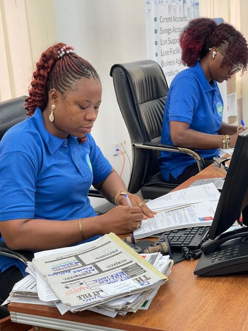 The image depicts two women in blue polo shirts engaged in administrative work at a desk. In front of the woman on the left, there is a bouquet arrangement of fresh flowers, including vibrant pink ros