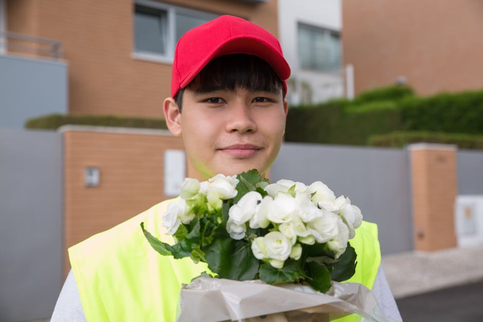 A young male flower delivery courier wearing a red cap and a high-visibility yellow vest is holding a bouquet of fresh white flowers, possibly freesias or similar blossoms, wrapped in white tissue pap