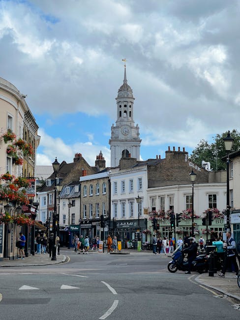 This image depicts a lively street scene in West Brompton Station SW5, with a historic clock tower featuring a spire rising above surrounding buildings under a partly cloudy sky. In the foreground, pe