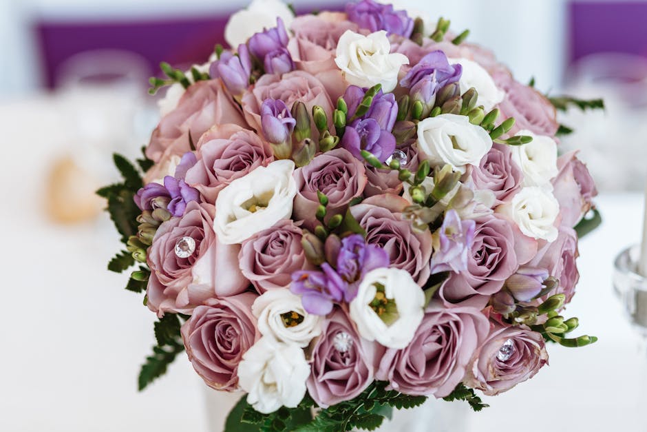 A close-up of a floral bouquet featuring pale pink roses, white lisianthus, and purple freesias, arranged in a round, lush style. The flowers are fresh with vibrant petals and green foliage, accented 